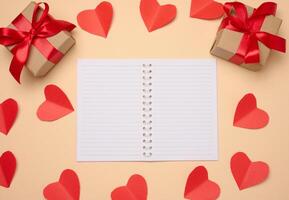 Open notebook with empty white sheets, red hearts and gift boxes on a beige background photo