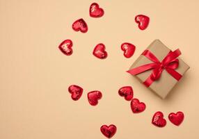 Box tied with a red ribbon and small hearts on a beige background, top view photo