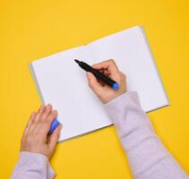 An open notebook with empty white sheets and a woman's hand holding a felt-tip pen on a yellow background photo