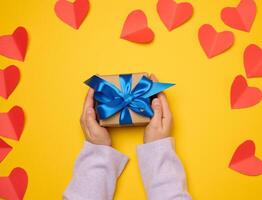 un mujer mano sostiene un regalo caja envuelto en un azul seda cinta en un amarillo antecedentes foto