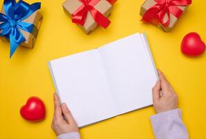 An open notebook with empty white sheets and gift boxes around on a yellow background, top view. Festive background for wish photo