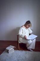 a medium-sized woman in light clothes reads a book while sitting in a white chair in a light room photo