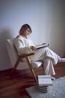 a medium-sized woman in light clothes reads a book while sitting in a white chair in a light room photo