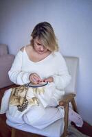 midsize woman in light clothes embroiders a picture in gold and beige tones while sitting on a white chair in a light room photo