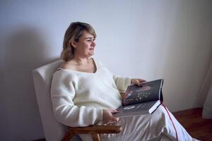 un talla media mujer en ligero ropa lee un libro mientras sentado en un blanco silla en un ligero habitación foto