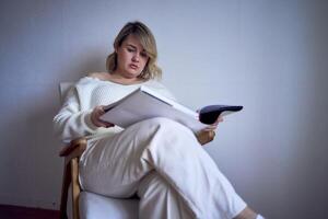 a medium-sized woman in light clothes reads a book while sitting in a white chair in a light room photo