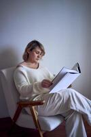 un talla media mujer en ligero ropa lee un libro mientras sentado en un blanco silla en un ligero habitación foto