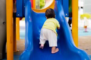 trasero de asiático bebé chico sube el para niños diapositiva en patio de juegos. niño jugando felizmente en verano o primavera veces. niñito jugando exterior. activo niño en vistoso deslizar. hijo Envejecido 1 año viejo. foto