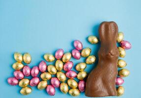 big milk chocolate bunny and pile of Easter candy eggs foil wrapped in pink and golden on blue background photo