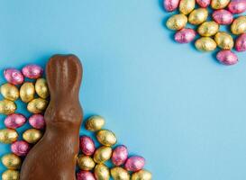 Milk chocolate Easter bunny and frame of pile of candy eggs wrapped in pink and golden foil on blue background photo