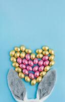 Easter bunny ears and heart shaped pile of candy eggs wrapped in pink and golden foil on blue background photo
