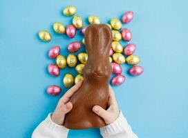 Child holding Easter chocolate bunny on blue background with candy eggs, Easter concept, top view photo