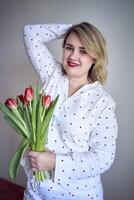 retrato de un hermosa talla media mujer en pijama con un ramo de flores de rojo tulipanes en un minimalista estilo foto