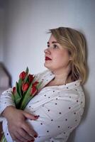 retrato de un hermosa talla media mujer en pijama con un ramo de flores de rojo tulipanes en un minimalista estilo foto