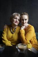 elegante antiguo madre y de edad mediana hija en amarillo suéteres abrazo y comodidad cada otro en un café foto