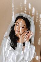 Beautiful, young, perfect model bride in a white lace dress sits in the room, bedroom in the morning, putting an earring on her ear. Wedding photography, portrait, concept. photo