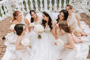 A brunette bride and her bridesmaids in matching cream dresses sit with the bride and rejoice with flowers in their hands near the ceremonial arch. Wedding in nature. photo