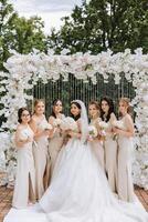 A brunette bride and her bridesmaids in matching cream dresses stand and rejoice with bouquets of flowers near the ceremonial arch. Wedding in nature photo