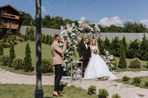 Wedding ceremony in nature. The bride and groom near the flower arch. The master of ceremonies at the wedding during the performance in the background of the bride and groom. photo