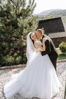 A handsome groom embraces his bride in a lush white dress and smiles in a beautiful outdoor setting. Under the open sky. High quality photo. A newlywed couple poses together on a sunny summer day. photo