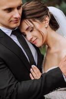 A handsome groom embraces his bride in a lush white dress and smiles in a beautiful outdoor setting. Under the open sky. High quality photo. A newlywed couple poses together on a sunny summer day. photo