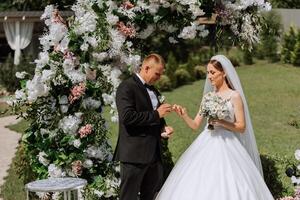 el novio pone en el de la novia Boda anillo durante el Boda ceremonia cerca el flor arco. verano Boda en naturaleza. ella dijo Sí. un conmovedor momento a el Boda ceremonia foto