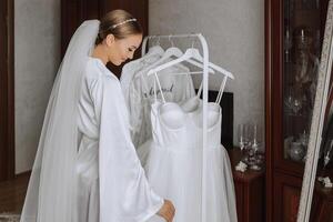 A beautiful bride with a long veil in her room, wearing a robe. Wedding dress on a mannequin. The bride in the morning before the wedding ceremony. photo