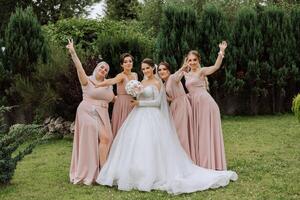 un morena novia y su damas de honor en idéntico rosado vestidos son en pie en contra el antecedentes de naturaleza. muchachas en idéntico vestidos son haciendo fuera a un boda. Boda en naturaleza. foto