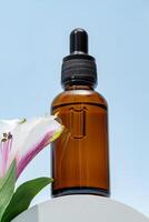 A brown dropper bottle of face oil displayed on a pedestal against a blue background with pink alstromeria flower. cosmetic package , unbranded mockup photo