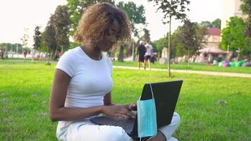 Young african american woman using laptop in park during covid 19 pandemic. Medical disposable mask hanging on display video