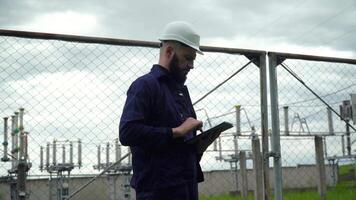 Engineer in special uniform standing with tablet near power station. Male monitoring system performance and making notes in program video