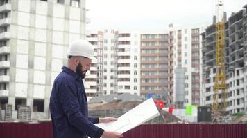 Portrait of a successful young engineer, architect, builder, businessman, wearing a white helmet, in a shirt, holding a project in his hand, a skyscraper background and a construction site video