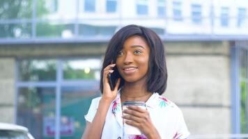volwassen Afrikaanse Amerikaans jong bedrijf vrouw is pratend Aan telefoon en drankjes koffie buiten in de buurt modern kantoor gebouw. einde van quarantaine video