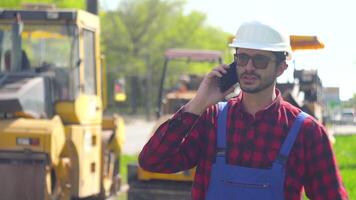 Baumeister im ein Helm und Uniform gegen das Hintergrund von ein Straße Konstruktion Seite? ˅ reden auf das Telefon video