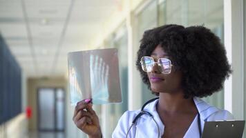 Female african-american doctor examines x-ray of leg, holding it in hands indoors. Specialist holds transparent image of leg in arms, and carefully researches it video