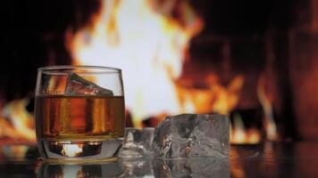 Glass of whiskey, scotch or bourbon with ice stands on a table against background of a fireplace with a flame. Alcoholic drink and home comfort concept. video