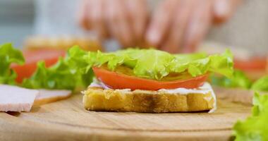 weiblich Koch im das Küche von das Haus sammelt ein Sandwich. Herstellung ein Sandwich mit Schinken, Salat, reif Tomate Scheiben und Stücke von Käse. schnell Essen Konzept. Ultra 4k video
