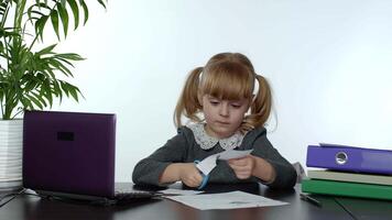 Child schoolgirl learns lessons at home sitting at table cutting with scissors shapes out of paper video