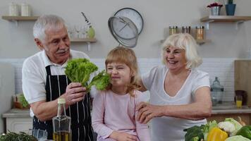 Happy vegan senior couple dancing with granddaughter child while cooking vegetables in kitchen video