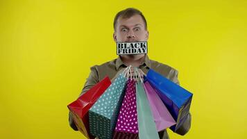 Surprised man showing shopping bags and Black Friday inscription in his mouth. Yellow background video