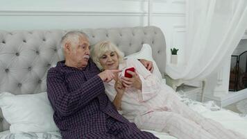 Senior elderly couple wearing pyjamas lying on bed looking on mobile phone laughing and having fun video