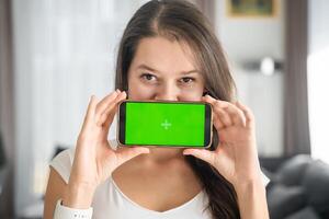 Young smiling woman shows smartphone with green screen in home living room closeup. Technology, gadget, connection. Advertising area, workspace mock up. High quality photo