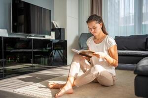 Young woman reads book sitting on the floor in the living room in sunny day photo