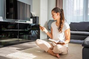 Young woman is trying to read book but is constantly distracted by smartphone and social networks photo