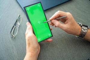 Close up view of man hand holding smart phone with green chroma key screen and stylus pen at his creative workspace photo