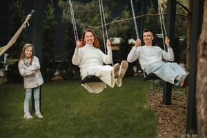 Mom and dad are riding on a swing and there is a daughter standing next to them. The family is resting on a swing photo