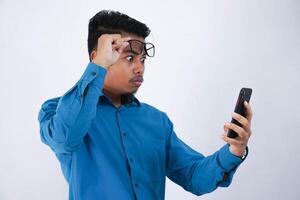 retrato de sorprendido y conmocionado hermoso joven asiático empresario con lentes en vistiendo azul camisa utilizando teléfono inteligente y tomando apagado lentes aislado en blanco antecedentes foto