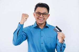 emocionado o contento joven asiático empresario con lentes participación crédito tarjeta con manos en puños vistiendo azul camisa aislado en blanco antecedentes foto