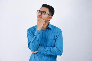 joven hermoso asiático hombre con lentes pensando y mirando para ideas mientras participación el barbilla vistiendo azul camisa aislado en blanco antecedentes foto