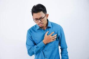 asiático joven hombre con lentes participación su cofre en cofre dolor o coronario corazón enfermedad vistiendo azul camisa aislado en blanco antecedentes foto